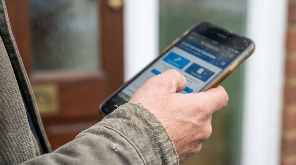 A tradesperson in work clothes standing at the front door of a UK terraced house, checking their phone to find a local service business website