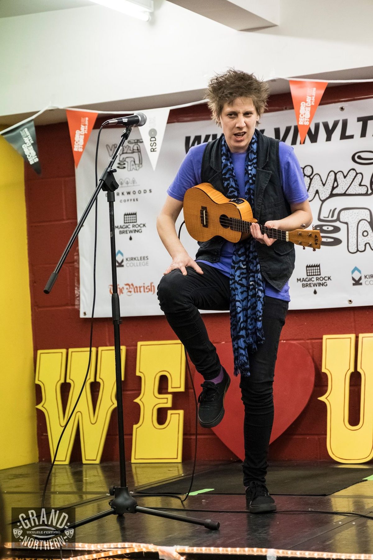 Fragile Lucy on stage, leg up, with her ukulele in front of large WE LOVE U letters