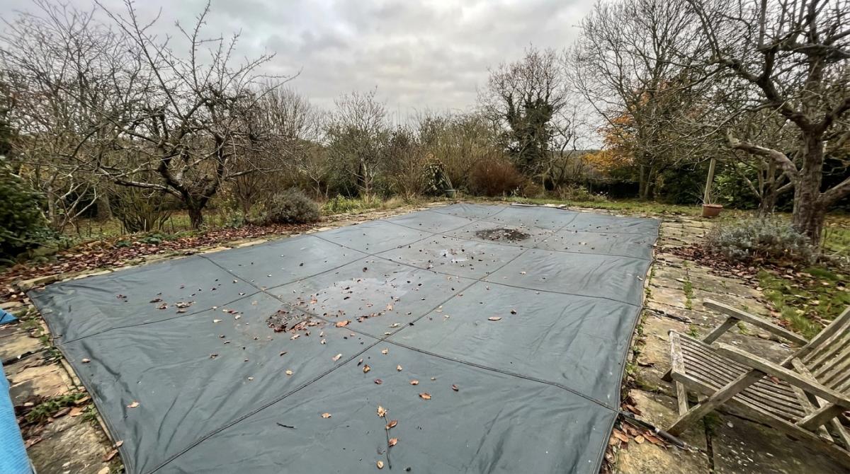 A residential outdoor swimming pool covered with a grey safety cover in an autumn garden, bare trees visible, the pool clearly unused and dominating the outdoor space