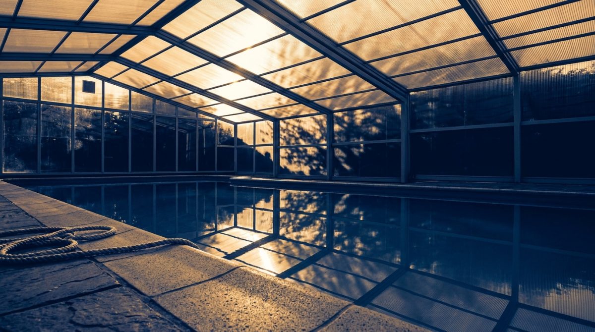 Interior of an aluminium and polycarbonate pool enclosure with dappled light filtering through the roof panels onto still blue water, viewed from poolside level